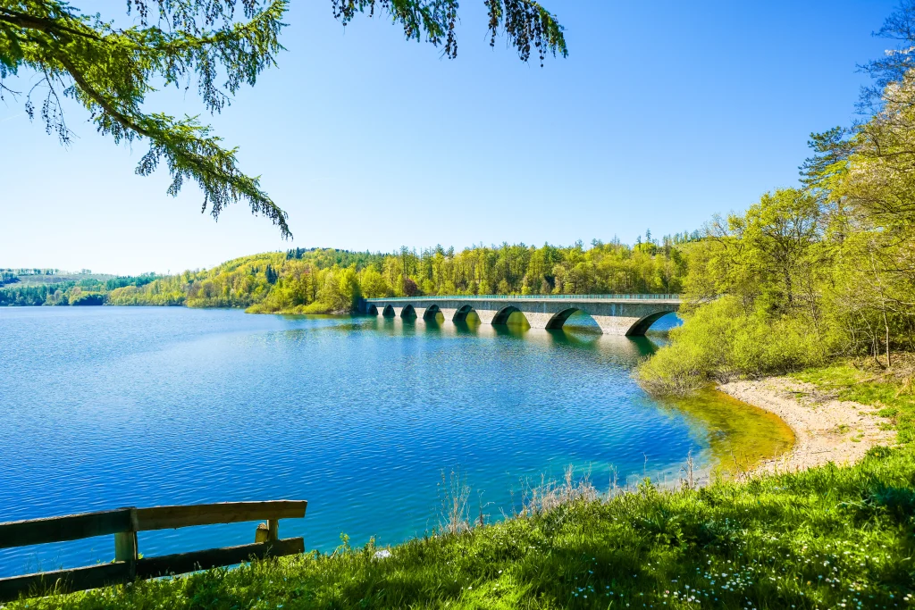 Steinerne Bogenbrücke über einen blauen Stausee im Sauerland, umgeben von grünen Wäldern und Wiesen